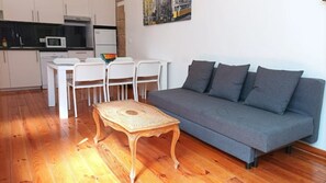 A modern living room with a grey sofa, wooden coffee table, and white chairs. A kitchen with white cabinets and a microwave is visible in the background.