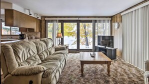 A living room with a sofa, a coffee table, a TV, and a view of trees through large windows. - Lakefront Eagle River Cottage w/ Private Dock (Eagle River)