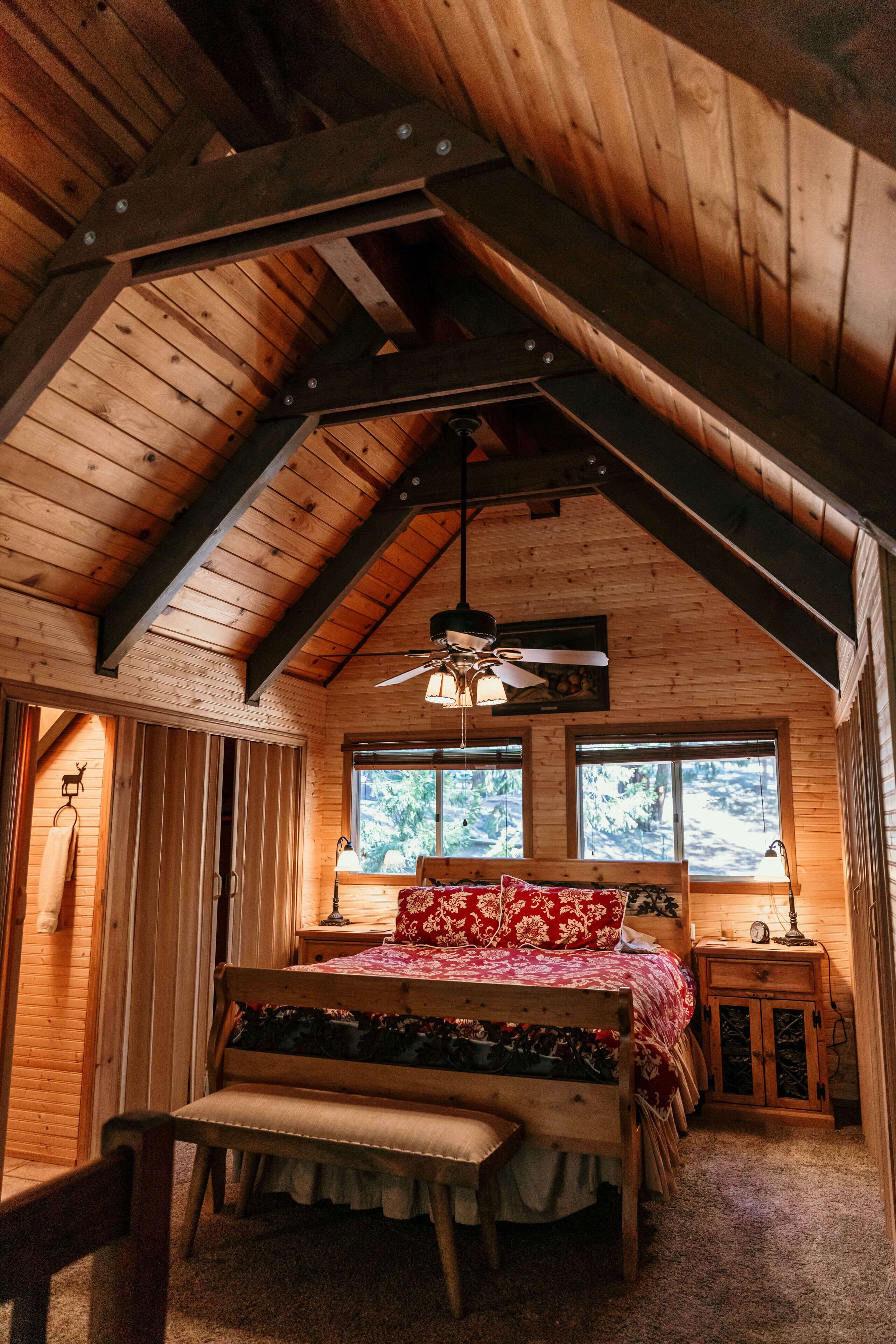 A wooden cabin bedroom with a sloped ceiling, a bed with floral bedding, a bench, a bedside table, and a window with a view of trees.