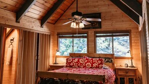 Un dormitorio de cabaña de madera con techo inclinado, una cama con ropa de cama floral, un banco, una mesita de noche y una ventana con vista a árboles.