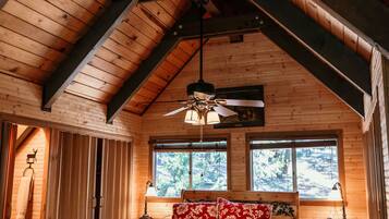 A wooden cabin bedroom with a sloped ceiling, a bed with floral bedding, a bench, a bedside table, and a window with a view of trees.