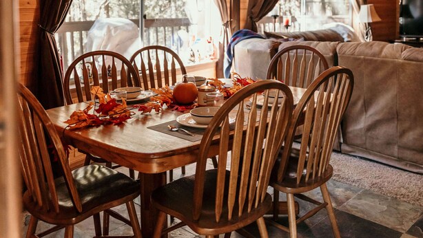 Una mesa de comedor de madera con sillas, un sofá y un televisor en una cabaña con ventanas amplias y techo de madera.