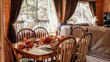 A wooden dining table with chairs, a sofa, and a TV in a cabin with large windows and a wooden ceiling.