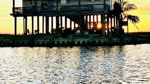 A two-story house with a balcony, situated on stilts over water, during sunset.