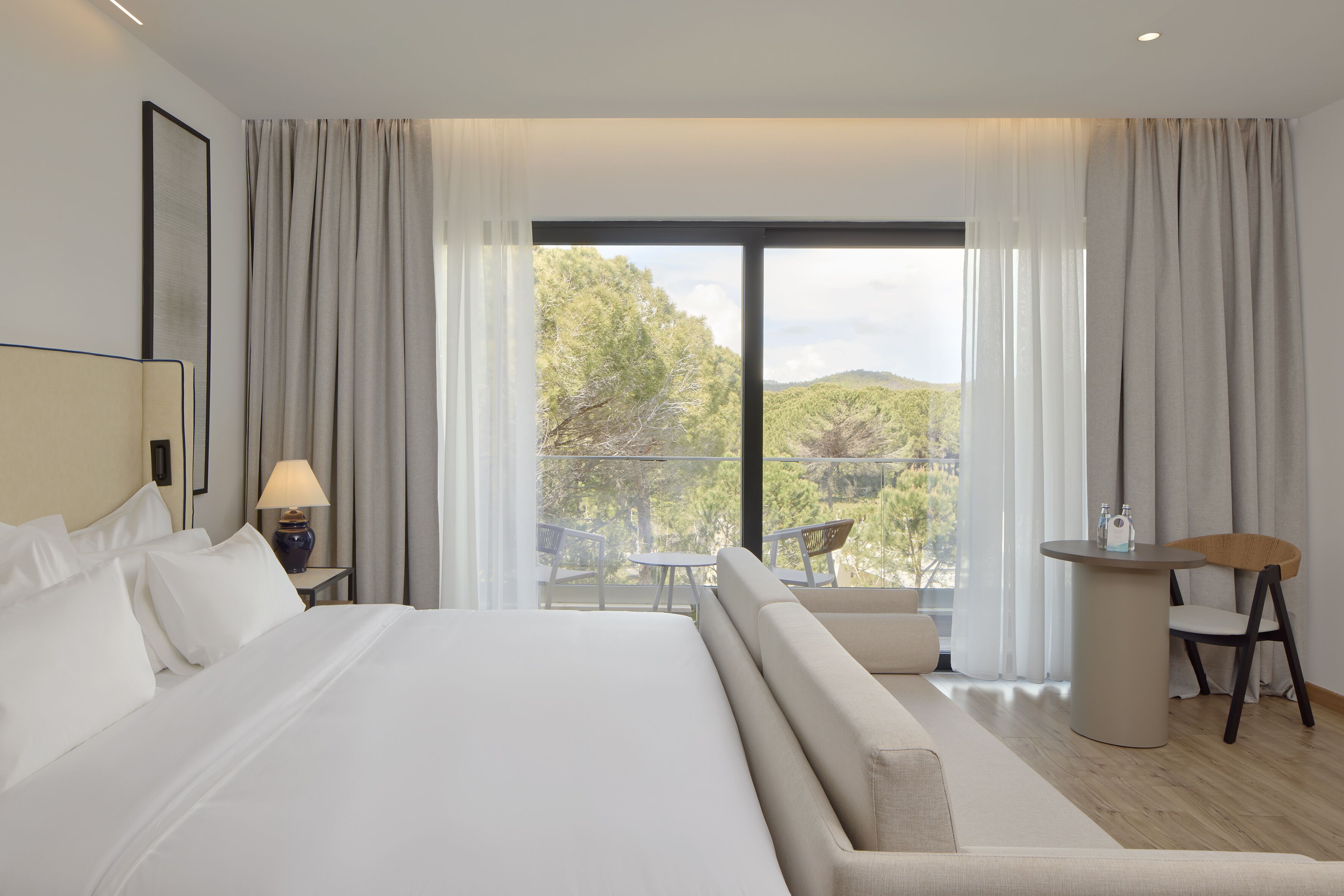 Habitación de hotel moderna con una cama grande, un balcón con mesa y sillas, y vistas al exterior.