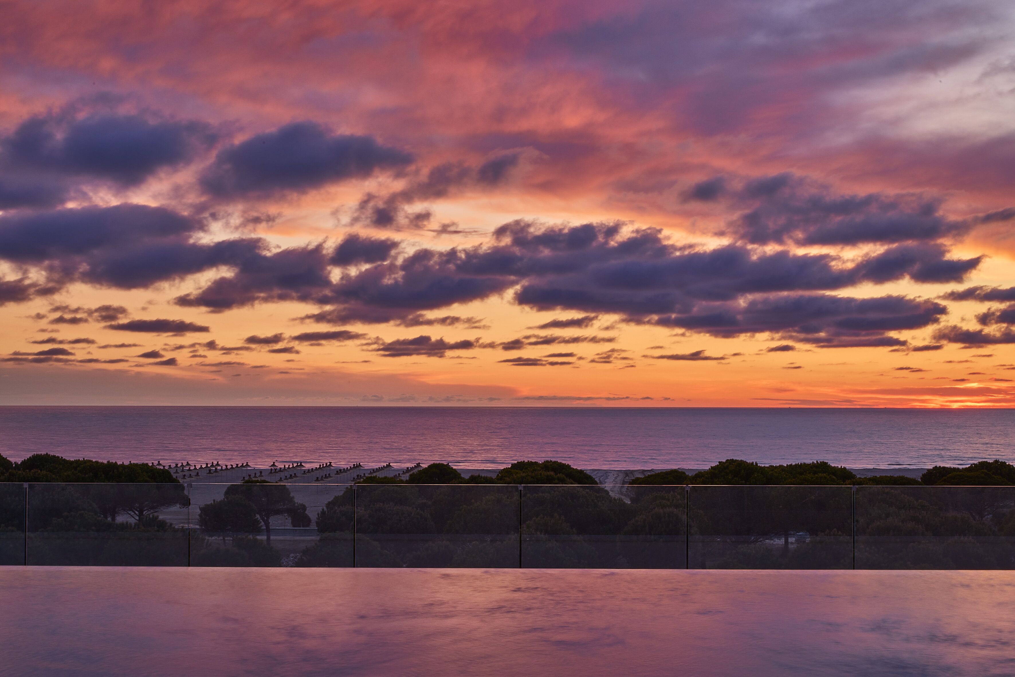 A sunset view from a balcony overlooking the sea, with a fence and trees in the foreground.