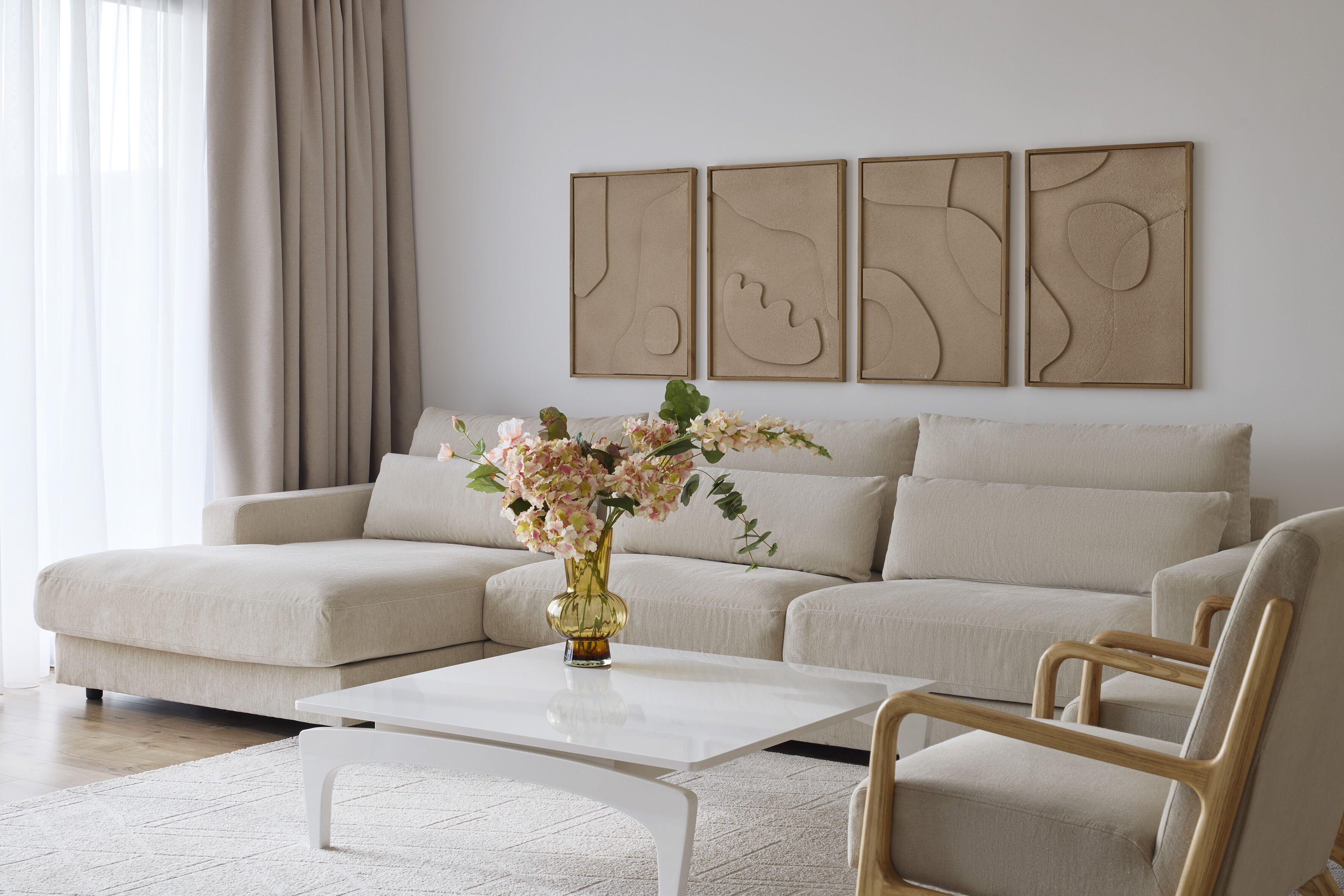 Un salon moderne avec un canapé d’angle beige, une table basse blanche et une chaise en bois. Des œuvres d’art abstraites sont accrochées au mur au-dessus du canapé.