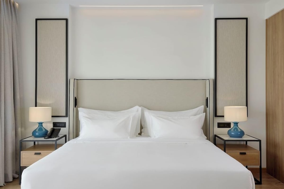 A hotel room with a large bed, two bedside tables with lamps, a nightstand, and a wall-mounted control panel.