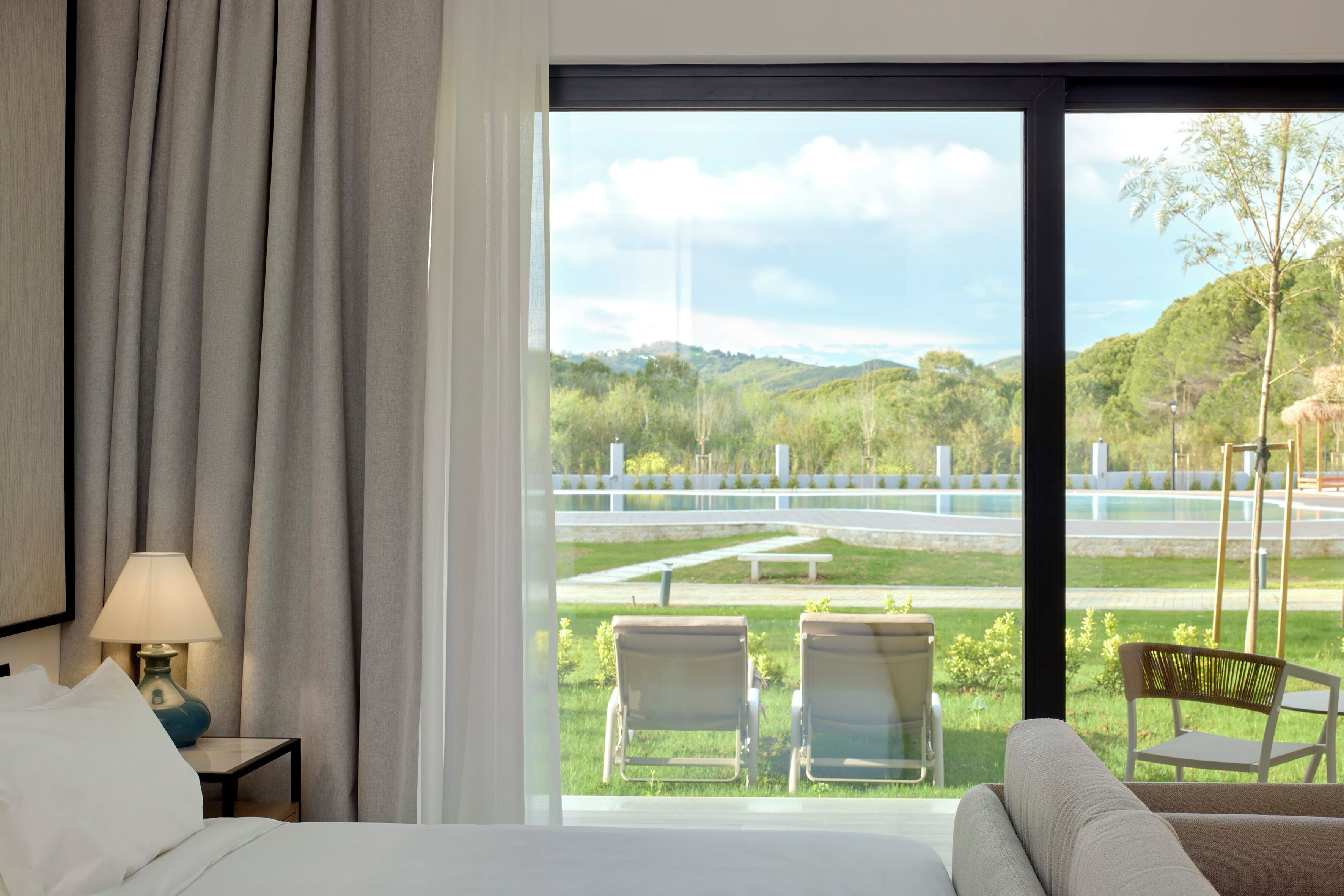 A hotel room with a large window, a bed, a nightstand with a lamp, and two chairs outside.