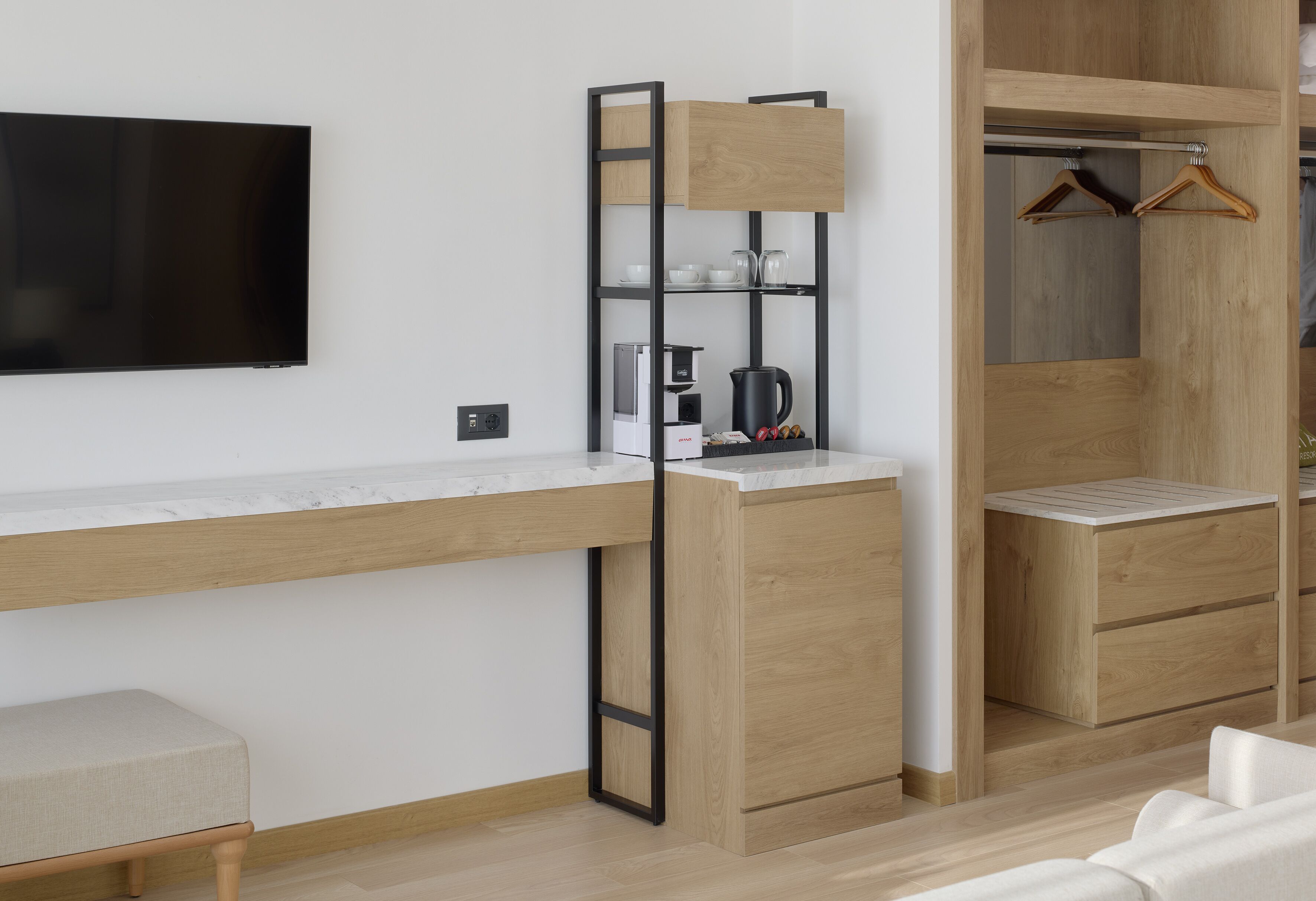 Habitación moderna con escritorio integrado, televisión de pantalla plana, cafetera y armario de madera con espacio para colgar.