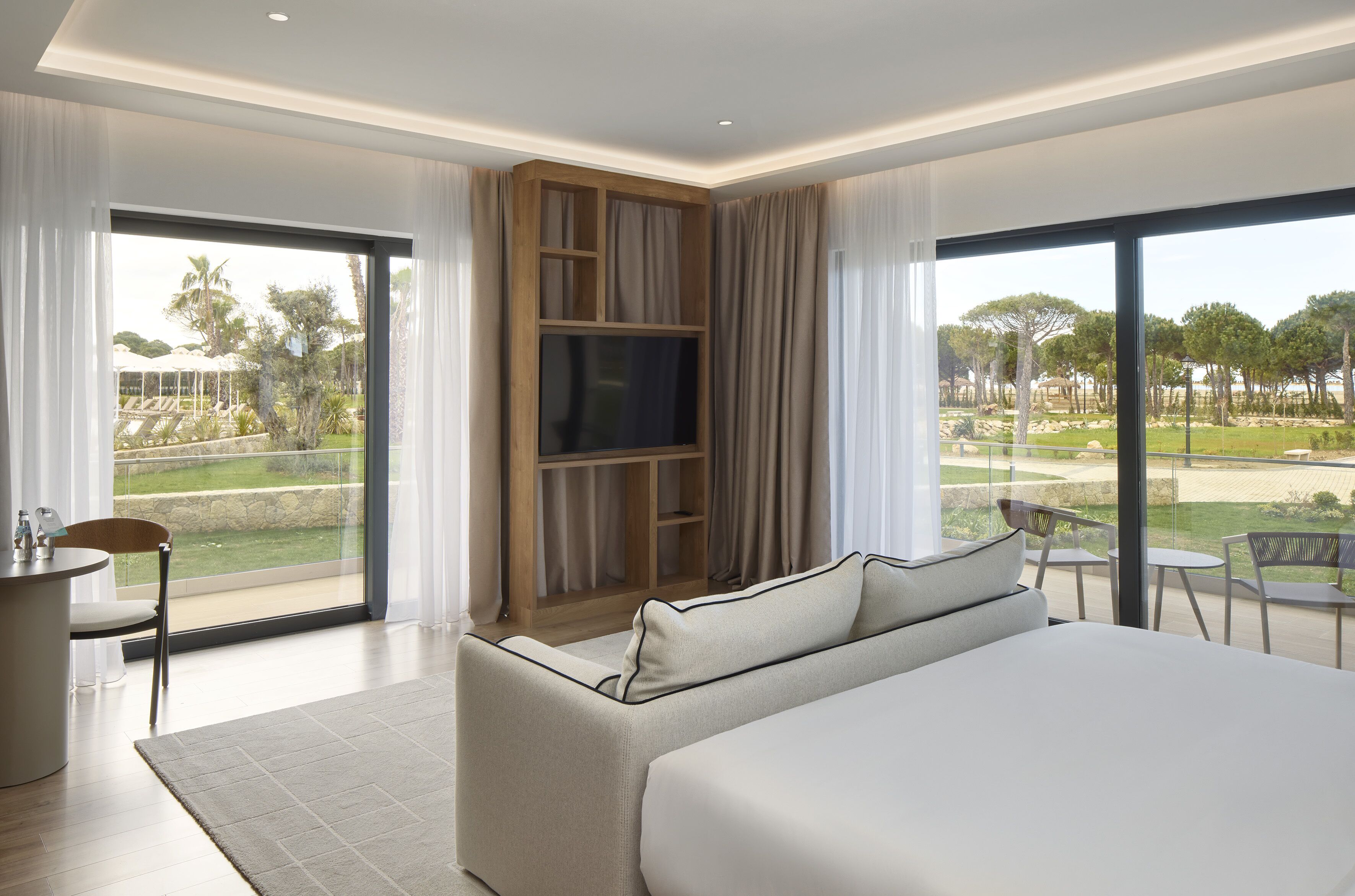 Ett modernt hotellrum med en stor säng, en tv som är monterad på en träpanel, en matplats med bord och stolar samt utsikt över en golfbana och träd.