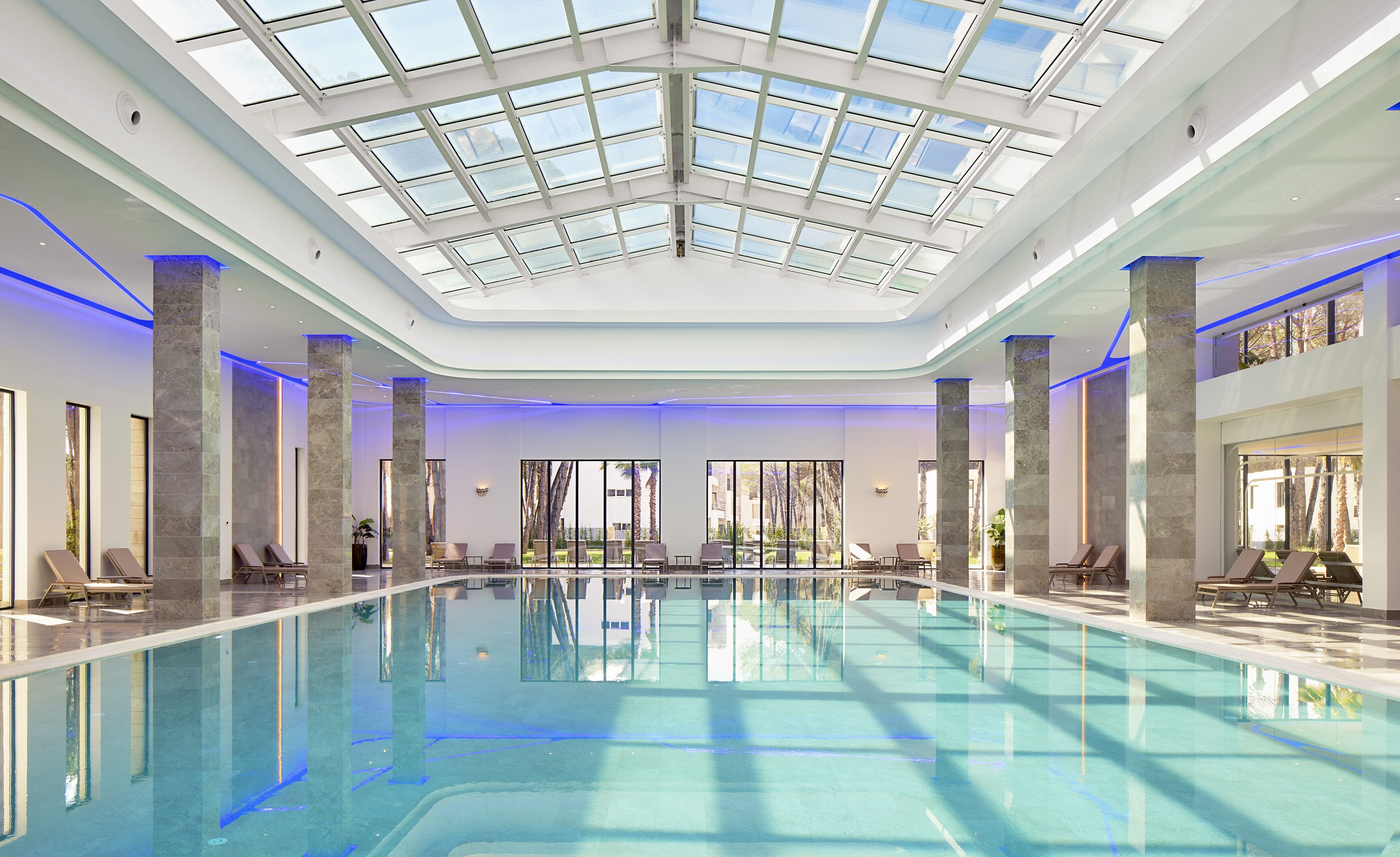 A large indoor swimming pool with a glass ceiling, marble pillars, and sunlit views of palm trees.