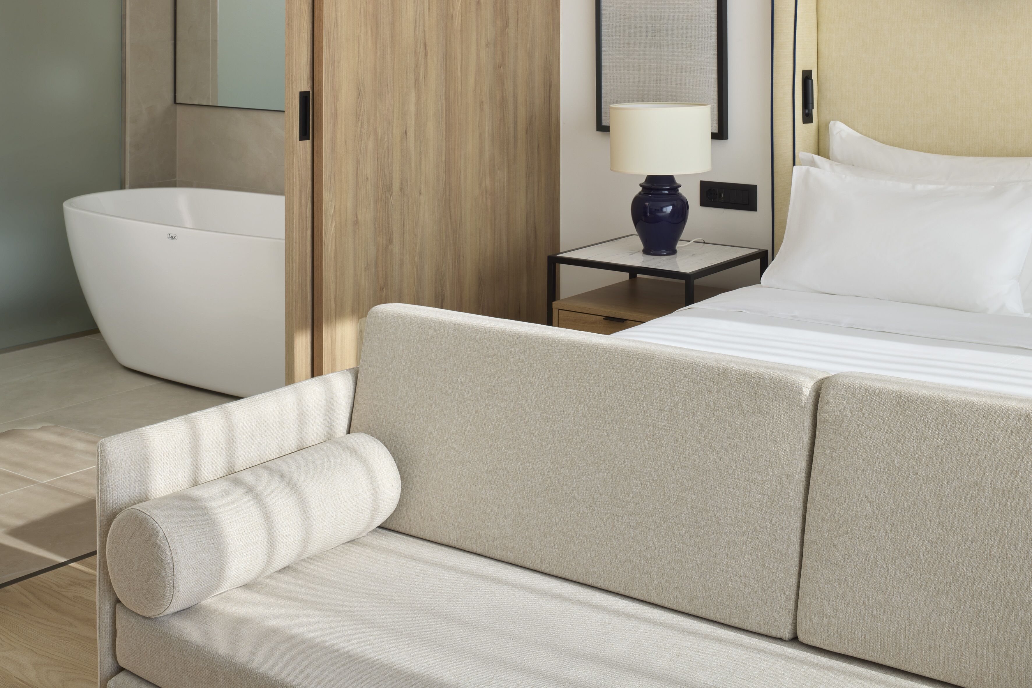 Ein modernes Hotelzimmer mit einer großen Badewanne, einem Sofa, einem Bett mit weißer Bettwäsche, einem Nachttisch mit einer Lampe und einer Wandpaneele aus Holz.