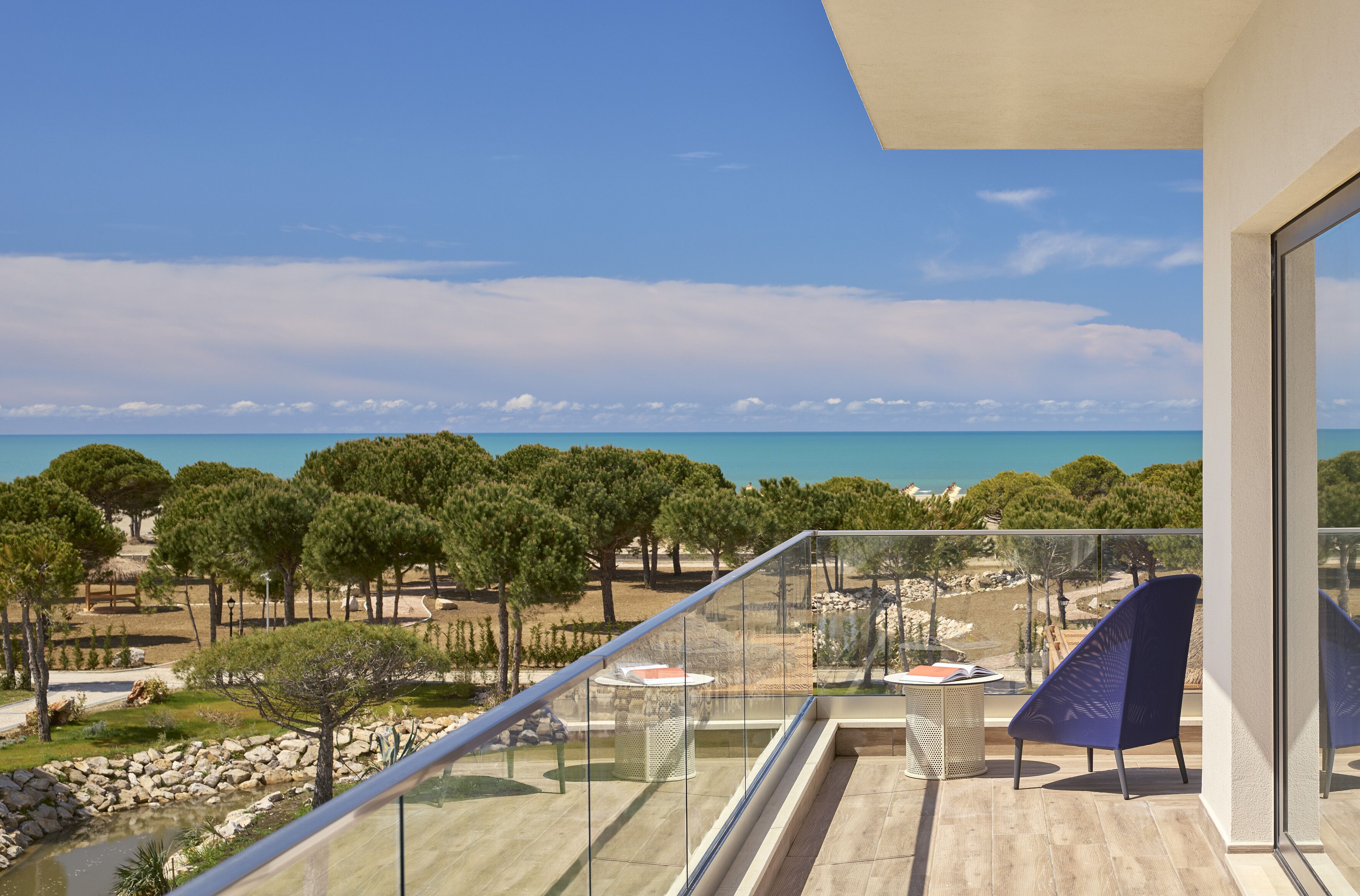 A balcony with a clear view of the sea, a grassy area with trees, and a modern building structure.