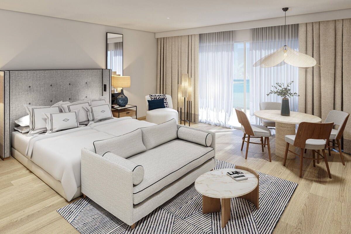 A modern hotel room with a large bed, a sofa, a dining table with chairs, and a large window with curtains.