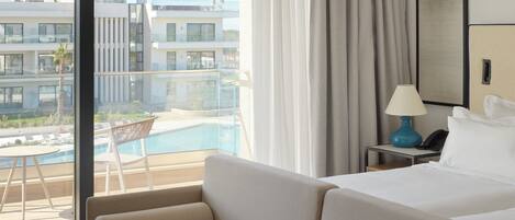A hotel room with a large bed, a nightstand with a lamp, a view of a swimming pool, and a balcony with chairs.