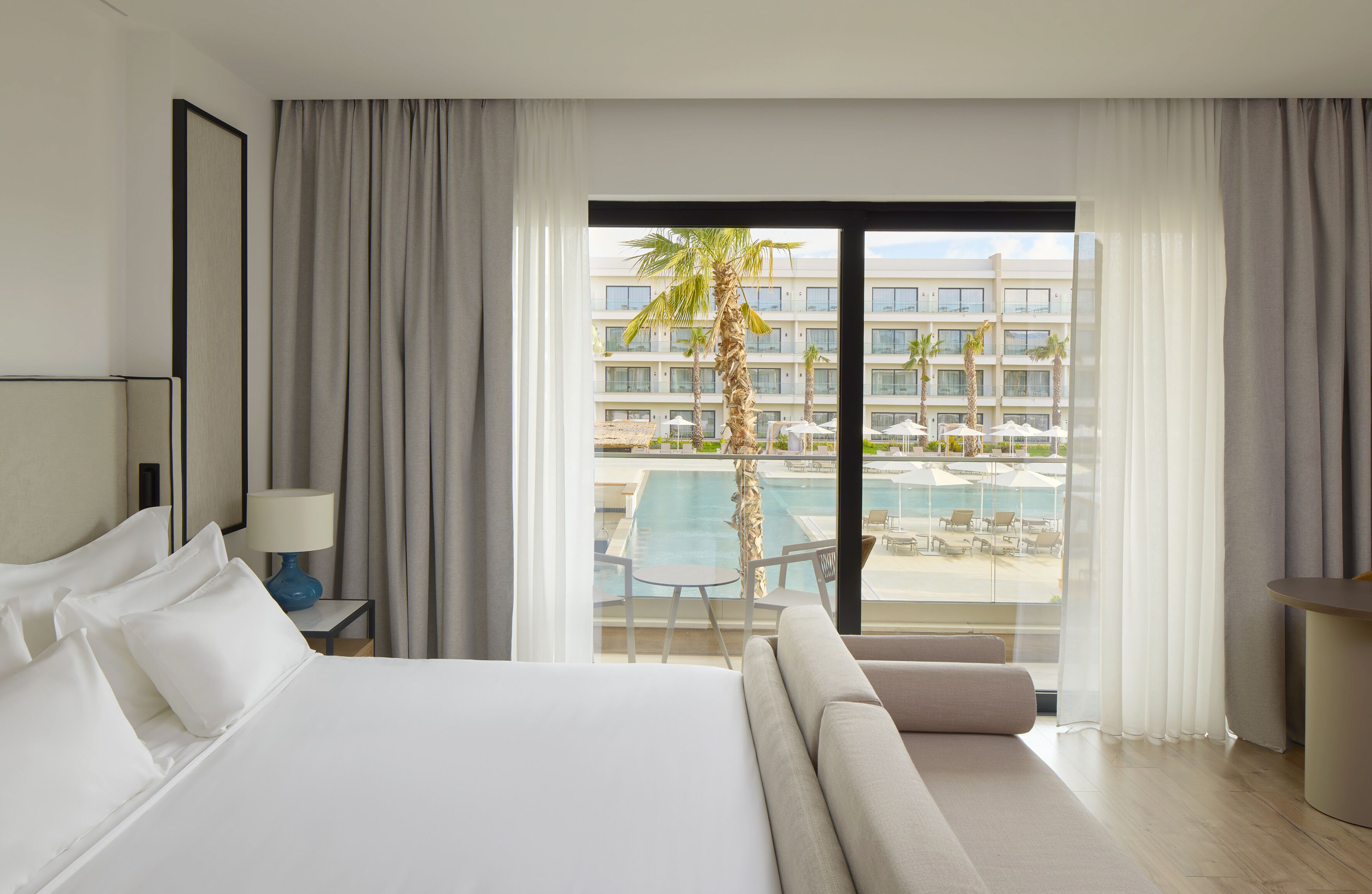 A hotel room with a bed, a nightstand with a lamp, a sofa, and a view of a pool and palm trees.