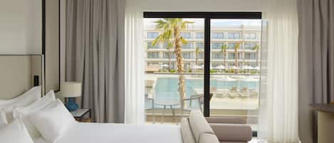 A hotel room with a bed, a nightstand with a lamp, a sofa, and a view of a pool and palm trees.