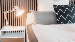 Ein ordentlich bezogenes Bett mit einer weiß-grauen Decke, einem schwarz-weißen Zickzack-Kissen und einer weißen Schreibtischlampe auf einem Nachttisch.