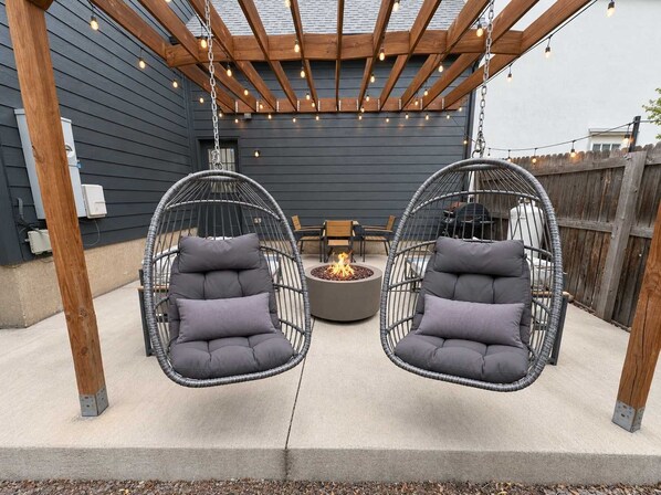 A patio with two hanging chairs, a round table, and a fire pit.