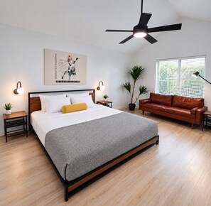 A modern bedroom with a bed, a ceiling fan, a brown leather sofa, and a window with blinds.