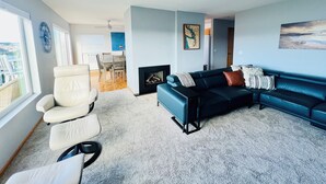 Een woonkamer met een bank, fauteuil en open haard. Achtergrond toont een eethoek met tafel en stoelen.