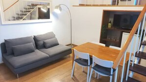Ein kompakter Wohnbereich mit grauem Sofa, einem Holzesstisch und zwei Stühlen. Eine Stehlampe ist vorhanden, und eine Treppe führt auf eine höhere Ebene.
