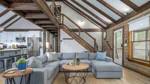 A spacious living area with a large grey sectional sofa, a round coffee table, and a wooden floor. A staircase with wooden beams leads to an upper level. - The Grande Lodge- Riverfront, Fiber, HotTub, Fish, DogOK, Game Room, Movie Room (Ellijay)