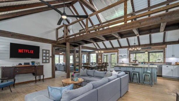 A spacious living area with a large gray sectional sofa, wooden beams on the ceiling, a kitchen with bar stools, and a flat-screen TV displaying 'NETFLIX'. - The Grande Lodge- Riverfront, Fiber, HotTub, Fish, DogOK, Game Room, Movie Room (Ellijay)