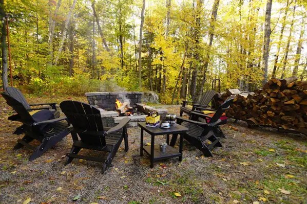An outdoor seating area with Adirondack chairs and a fire pit surrounded by a forest.