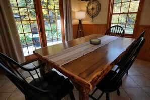 A wooden dining table with a placemat, four black chairs, a wall clock, and large windows with a view of greenery.