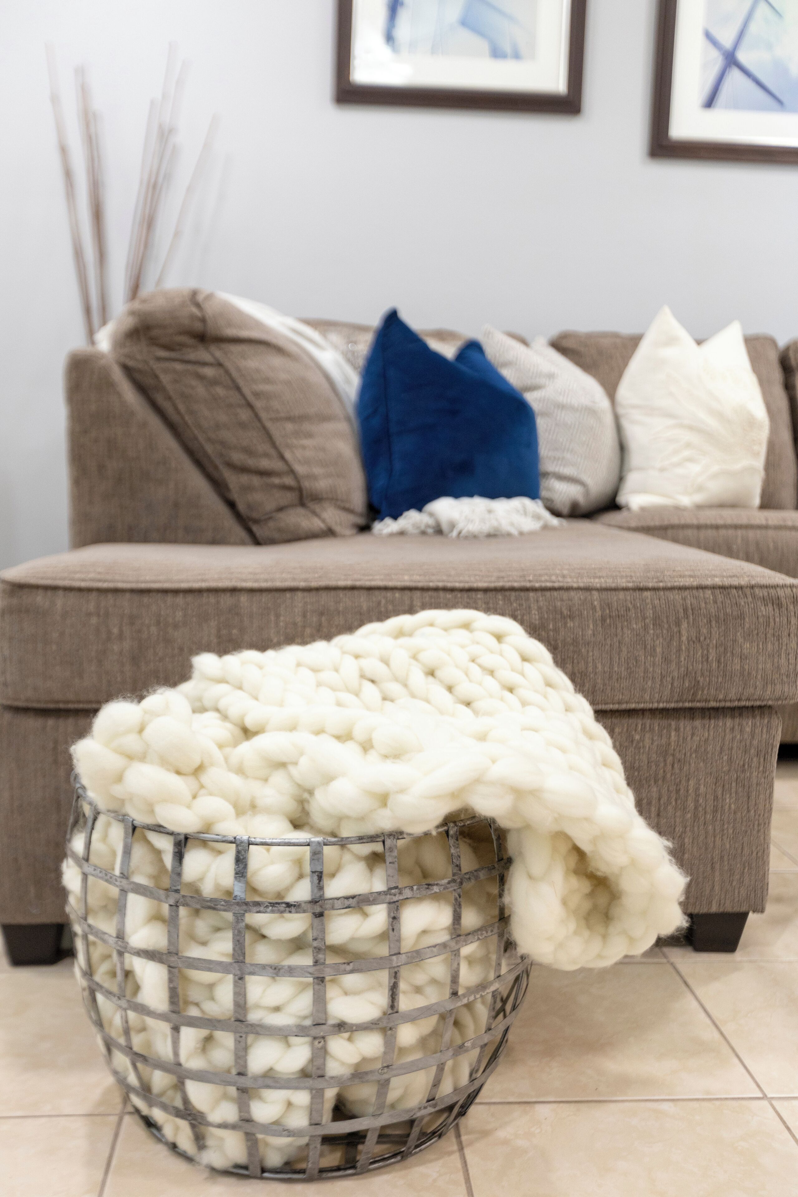 Ein Wohnzimmer mit grauem Sofa, weißer Strickdecke und einem Metallkorb voller weißer Wolle.