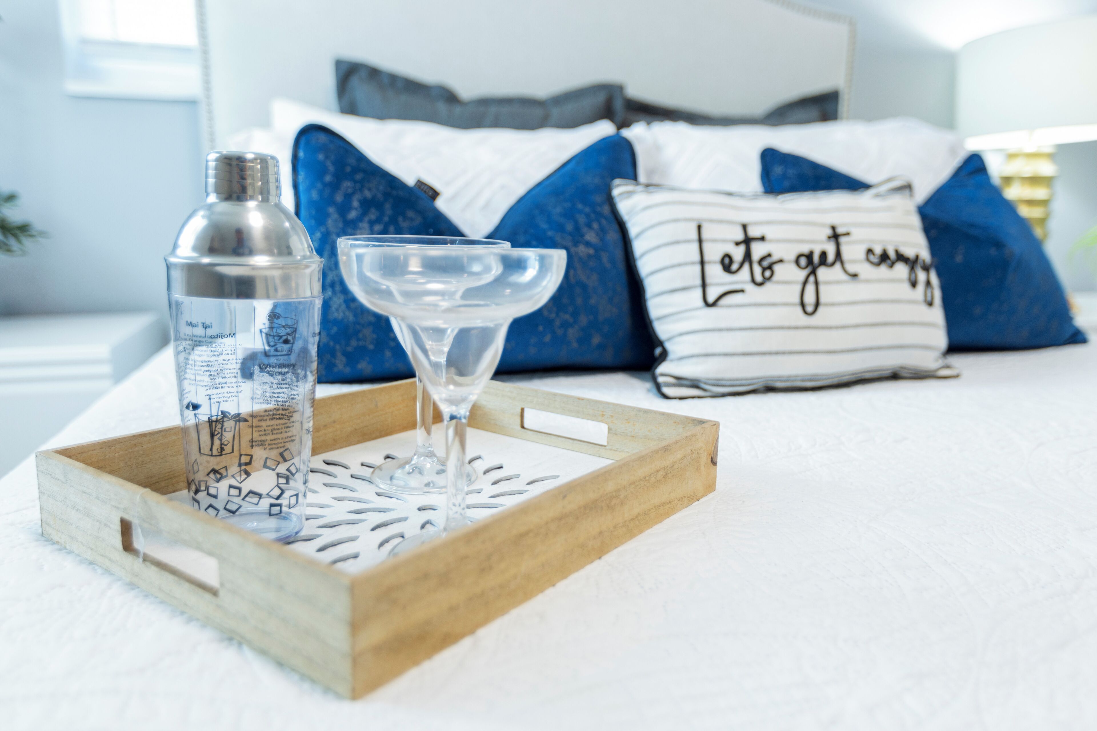 A wooden tray with a bottle and a glass on a bed with blue and white pillows, one with a handwritten message.