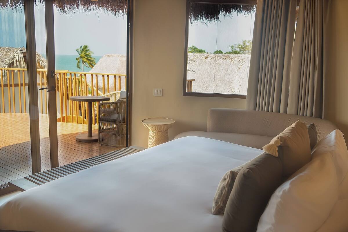 A hotel room with a large bed, a small table, a chair, and a view of the beach through a sliding door.