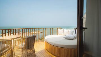 Een rond bed met wit bedgoed, een houten terras, rieten stoelen en uitzicht op zee.