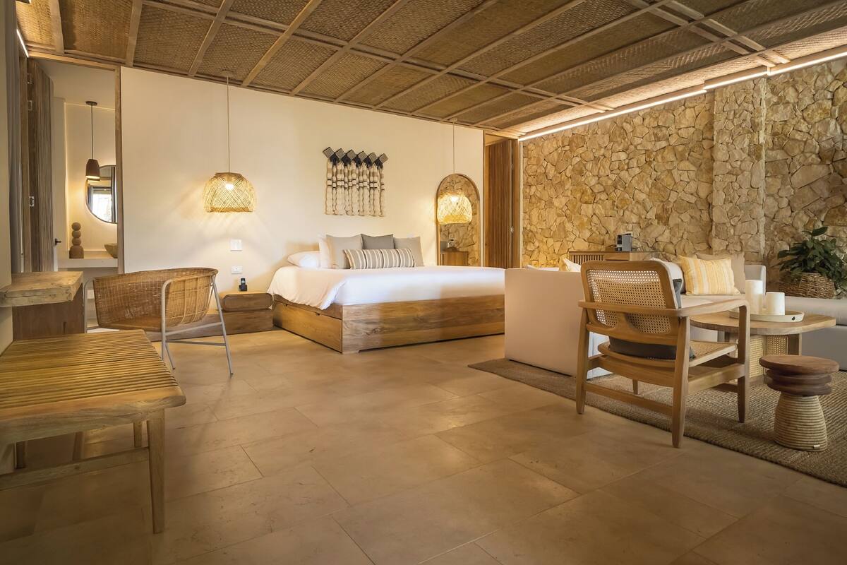 A bedroom with a bed, bedside tables, a chair, a small table, and a stone wall.