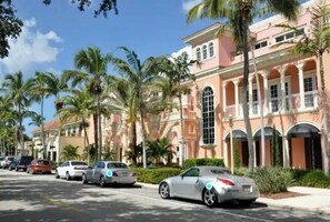 A street view with a row of parked cars, palm trees, and a pink building with arched windows and columns.