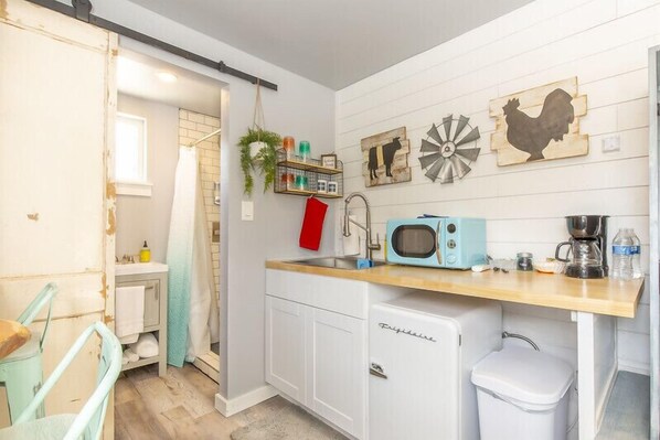 A compact kitchen with a microwave, coffee maker, and a small refrigerator. A red towel hangs on the door, and a shelf with various items is mounted on the wall. - The Container Retreat @ 290 Wine Trail #2 (Blanco County)