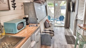 Une cuisine compacte équipée d’un four à micro-ondes, d’une machine à café et d’un petit évier. On y trouve également un canapé avec des coussins et une porte donnant sur un espace extérieur aménagé pour se détendre.