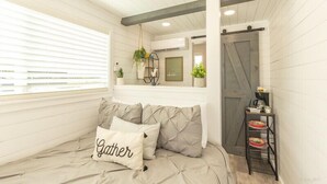 Un dormitorio acogedor con cama, almohadas, una mesita de noche, una ventana con persianas y una puerta.