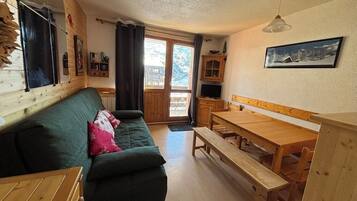 Une pièce chaleureuse avec un canapé vert, une table en bois et un banc. On y trouve une fenêtre avec des rideaux, un téléviseur et un tableau encadré au mur.