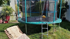 Een trampoline met veiligheidsnet, omgeven door een gazon en een houten terras.