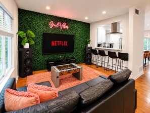 A modern living room with a flat-screen TV, a foosball table, a sofa, and a kitchen area with bar stools.