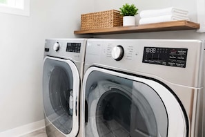 Une buanderie moderne équipée de deux machines à laver à chargement frontal, d’un panneau de commande et d’une étagère où sont rangées des serviettes et une plante.