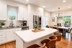 A modern kitchen with white cabinetry, stainless steel appliances, a central island with bar stools, and a dining area with a wooden table and chairs.