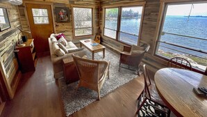 Una cabaña con interior de madera, un sofá, una mesa de comedor, sillas de mimbre y grandes ventanales con vistas a un lago.