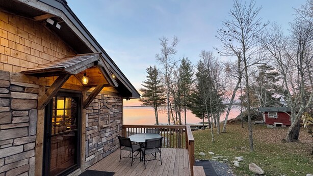 Una cabaña de madera con fachada de piedra, una terraza con mesa y sillas, y vista a un lago y árboles.