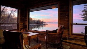 Interior acogedor de una cabaña con un gran ventanal que ofrece vistas a un lago tranquilo y árboles al atardecer.