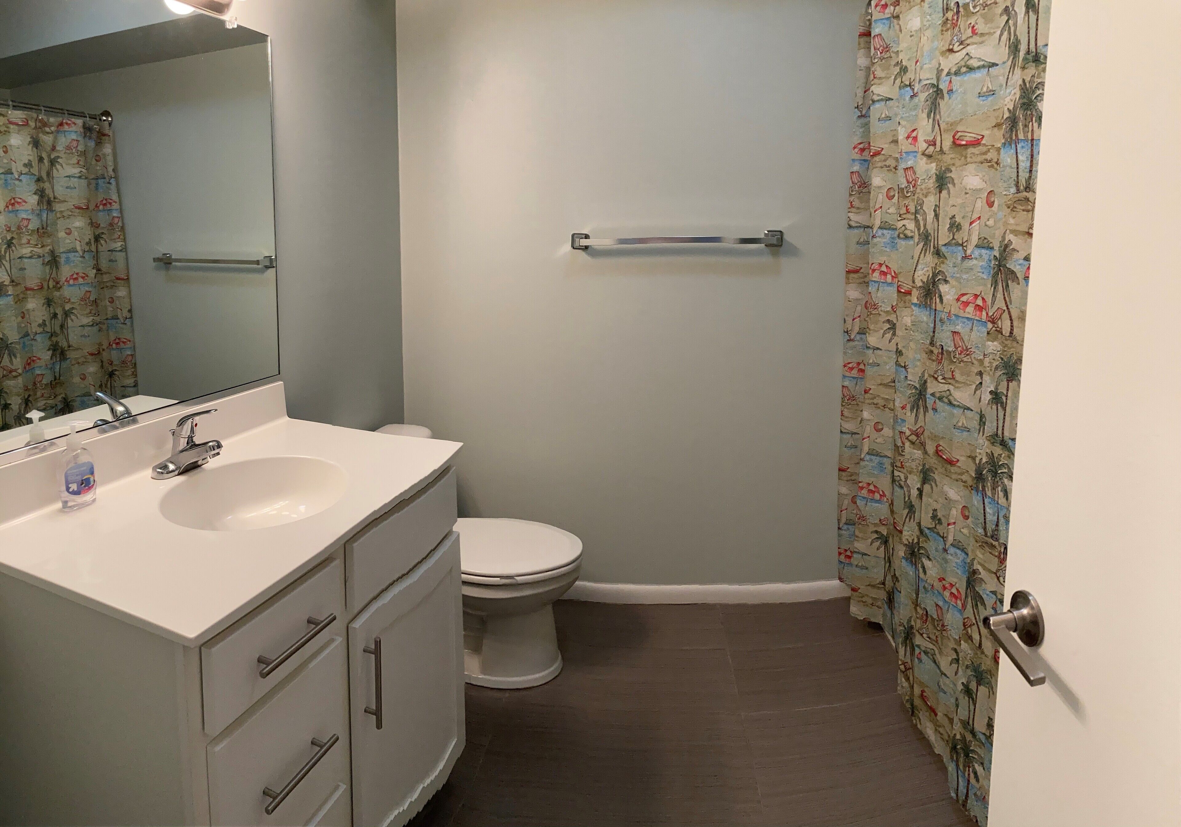 Une salle de bain avec un lavabo blanc, un miroir et des toilettes. On trouve un porte-serviettes au-dessus des toilettes et un rideau de douche orné d’un motif de plage.
