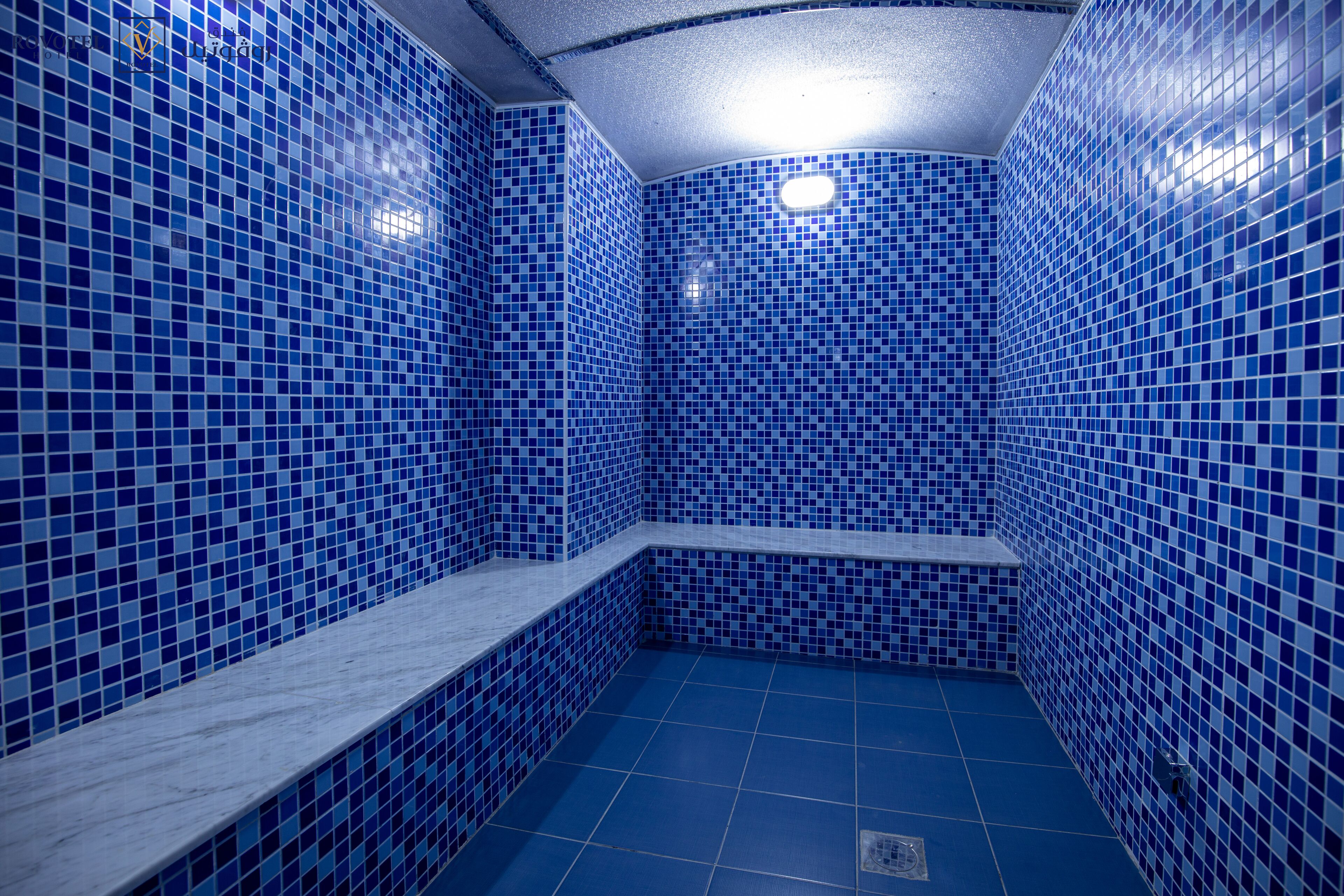 A bathroom with blue-tiled walls and floor, a white bench, and a visible drain.